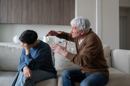 Mad aggressive senior man yelling and gesturing at crying stressed wife during family conflict, angry elderly husband in breakdown shouting at devastated woman sitting on couch covering face with hand
