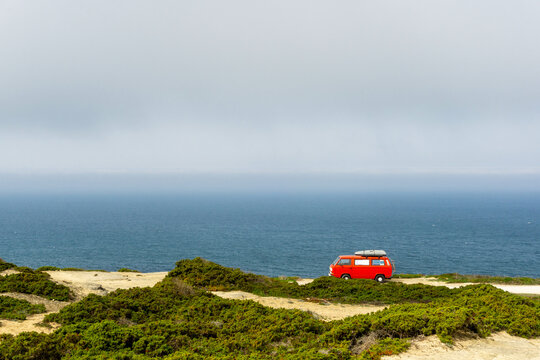 Red vintage camper van with a surfboard on the roof, parked on a cliff overlooking the vast blue Atlantic Ocean under a hazy sky, symbolizing freedom and adventure