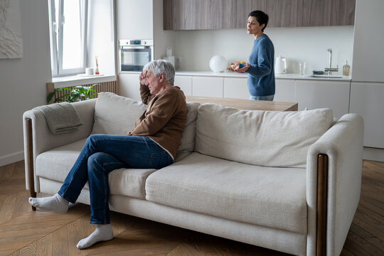Frustrated elderly husband sitting on sofa in room listening while complaining wife arguing at back, senior marriage crisis and domestic argument, aged couple in family quarrel and confrontation.