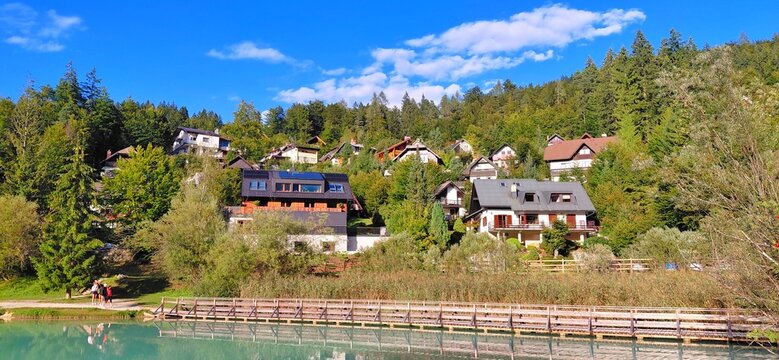 Scenic view of Lake Jasna in Kranjska Gora, Slovenia. Alpine chalets on a green forested hill reflecting in the crystal clear turquoise water with a wooden bridge under a blue sky. Summer travel.
