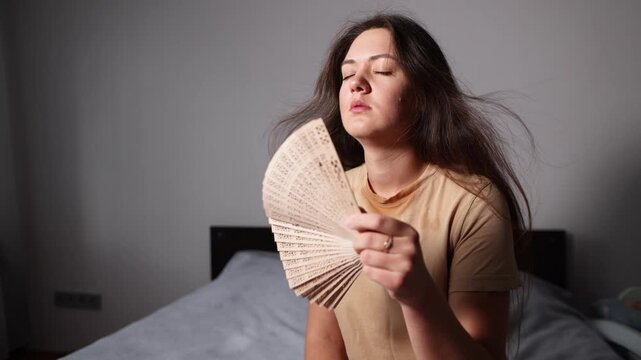 Young woman fans herself on bed while sweating heavily in extreme heat. Blurred baby crib suggests a mother coping without air conditioning.