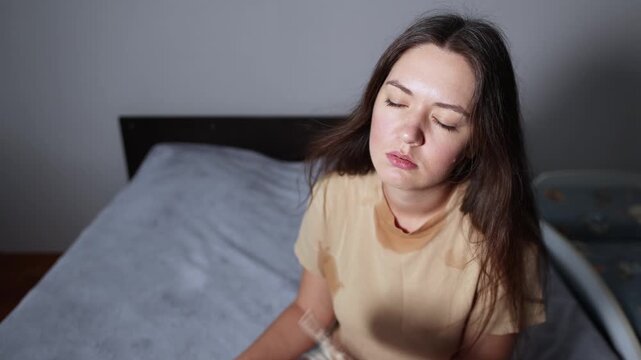 Young woman sits beside baby crib and fans herself with hand fan. Slow motion footage shows visible sweating and fatigue during extreme heat.