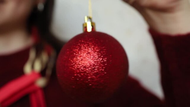 Young woman brunette in festive Christmas look keeping in hand and playing with red sparkling ball ornament. Symbol of celebration, Christmas holidays, New year. Sparkling vibe and party mood