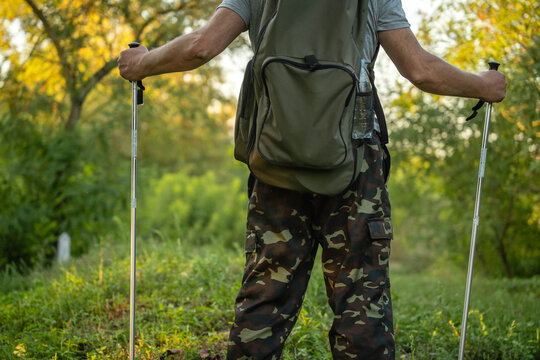 Backpack hiker pauses on forest path, gripping trekking poles with both hands. Camouflage trousers and green pack suit active outdoor walk, while relaxed posture suggests calm focus in warm sunlight.