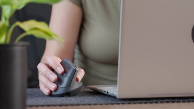 Closeup of woman using vertical ergonomic mouse beside laptop on desk. Computer work with ergonomic equipment in home office