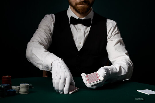 Casino gambling and professional gaming, dealer in tuxedo and white gloves shuffling playing cards at a green felt table with poker chips in dark room