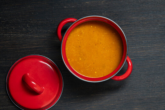 Homemade pumpkin soup in a red pot. Top view of delicious hot orange cream soup in vibrant red enamel pot with an open lid, served on wooden table. Perfect autumn or winter comfort food concept