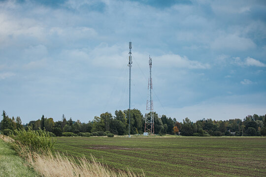 Communication towers rising above cultivated farmland and meadow grasses beneath a cloudy blue sky, with trees forming a rural horizon and open countryside landscape