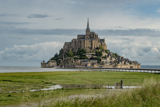 Road to Mont-Saint-Michel during high tide with rising water, tidal phenomenon on Atlantic's ocean coast, view on UNESCO heritage site and iconic Normandy landmark and green pastures