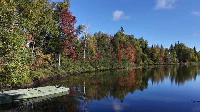 Drone footage flies over the mirror-like waters of Lac du Quinzi&egrave;me Mille, passing near rowboats along the shore with colorful autumn forest reflected in the water near Saint-Mo&iuml;se, Quebec.