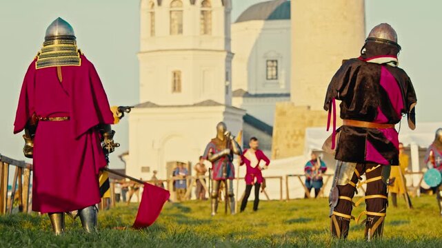 Armored knights prepare for battle near white stone temple. Historic stone church towers over warriors standing on green grass while waiting for medieval tournament combat.