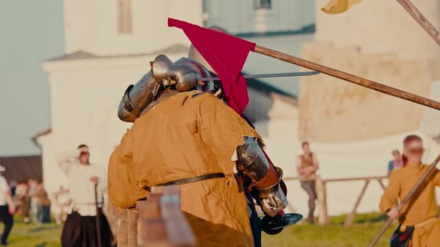 Marshal uses red flag to separate fighting knights. Official pushes bright fabric between armored warriors stopping intense historical medieval battle near wooden fence.