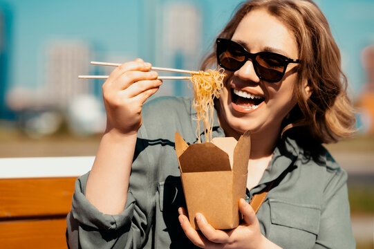 Delicious noodles from box with chopsticks &mdash; street food choice. Laughing joyful woman in sunglasses having outdoor lunch break on sunny street. Asian fast food and ready to eat wok concept.
