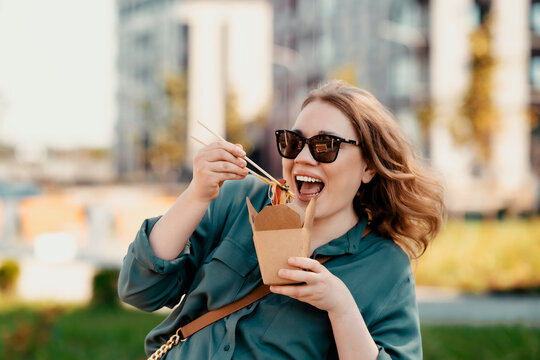 Fast food &mdash; tasty Asian noodles for meal. Smiling casual woman in sunglasses enjoying street food from paper carton box using chopsticks. Urban city lifestyle and ready to eat lunch break.