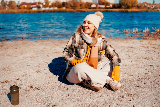 Positive woman in autumn park resting near river &mdash; healthy lifestyle choice. Smiling traveler sitting on sand exploring nature. Relaxing weekend walk bringing amazing psychological joy.