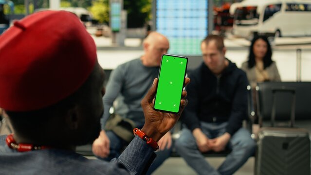 Black man holds a vertical chroma key phone in a busy bus station while passengers wait for an intercity coach; the mockup screen appears in a crowded terminal.