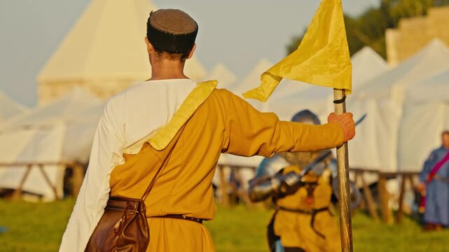 Medium shot shows marshal holding yellow flag at medieval tournament. Blurred armored knights clash with weapons in background during sunny historic reenactment festival on green grass.