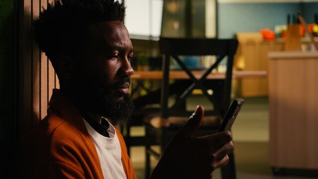 African American man uses a mobile phone indoors for social media and texting in a cozy home environment with a warm, relaxed atmosphere.