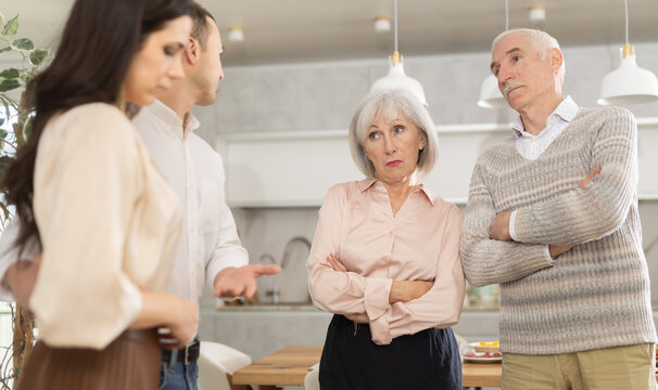 Young adult man presenting modest girlfriend to elderly parents, expressing disapproval and concern during tense meeting in family home