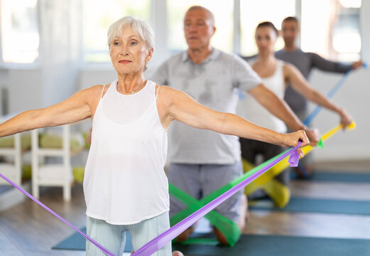 Elderly woman performs exercises with elastic band during Pilates strength training, kneeling. Fixation of tape under knee, tension of tape with help of hand strength, circular strength training