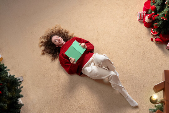 cheerful woman lying on the floor holding a book near a decorated Christmas tree soft carpet background empty room copy space