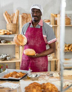 Smiling experienced black baker holding golden loaves of crusty bread, proudly presenting freshly baked goods in cozy private bakery environment..