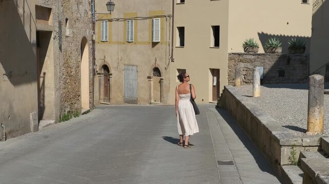 Woman in white dress walking down an old italian street. Young woman in a summer dress walking alone through a deserted street of an ancient tuscan village, enjoying her vacation
