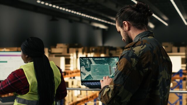 Adult man soldier supervising operational activities in military base, reviewing real-time surveillance systems on laptop interface; supply chain management and warehouse oversight.