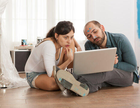 Paint-splattered couple focuses on a laptop during a renovation break, thoughtfully reviewing options for their DIY room makeover and decorating project.