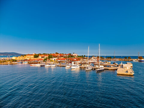 Moored sailing yachts beside concrete marina pontoons. Tall masts rise in front of waterfront buildings with orange roofs beneath a clear blue sky.
