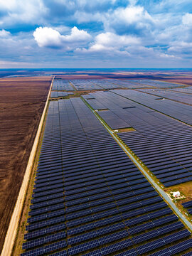 Solar farm boundary stretching toward the horizon in vertical view. A straight dirt road separates brown farmland from parallel arrays beneath white and gray clouds.