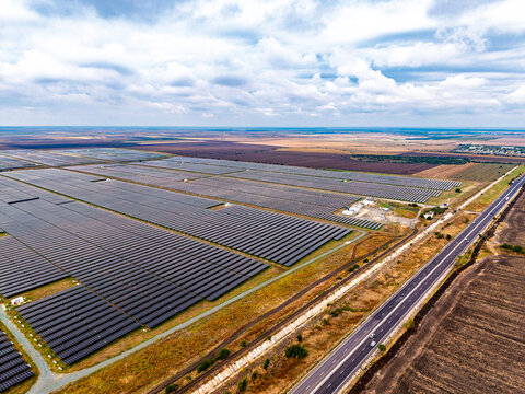 Aerial solar farm view with a highway along the right edge. Rows of dark panels surround an equipment compound under scattered clouds across a flat landscape.