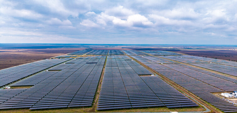 Panoramic view of photovoltaic rows extending toward the horizon. Straight service lanes divide dark solar modules beneath layered clouds across open farmland.