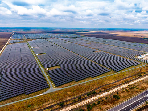 Solar installation, aerial overview of photovoltaic fields near highway. Countless dark energy panels stretch across vast terrain beside an asphalt road and railway track.
