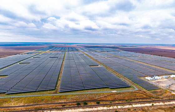 Solar park, aerial view of massive photovoltaic grid and rural fields. Structured rows of dark energy panels extend across open rural land beneath an overcast sky.