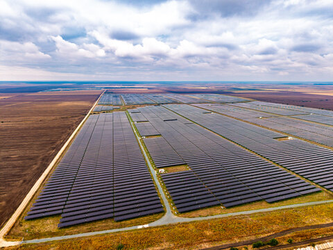 Solar power station, aerial perspective of photovoltaic modules on plains. Endless rectangular collector blocks span across dry agricultural land under heavy cloud cover.