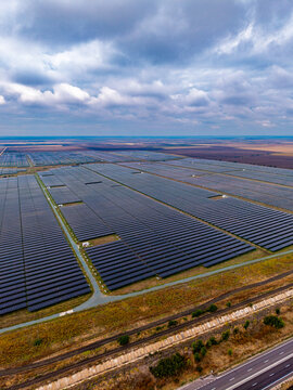Vertical solar farm view with a highway in the foreground. Rectangular panel blocks extend across farmland under gray clouds, with service roads between arrays.