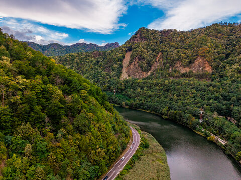 River bend with a road below rocky forested ridges. Green trees cover both banks beneath broken clouds, with vehicles following the near shore.
