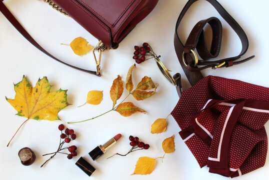 Lifestyle. A burgundy bag and scarf, a brown leather belt, and lipstick on a white background with autumn leaves, red berries, and chestnuts. Beauty and fashion. Top view, copy space, background, spac