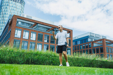 Fototapeta premium Man walks on grass while holding a smartphone near modern buildings during daytime