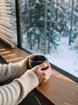 Woman holding a steaming ceramic mug of hot tea by a large window overlooking a scenic snowy pine forest