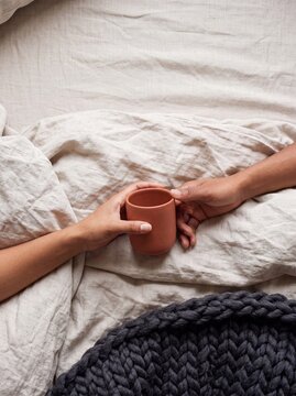 Top view of a couple holding a terracotta ceramic mug together in bed surrounded by cozy textured linen sheets and a knitted blanket