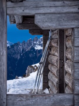 Pair of cross country skis and poles leaning against a rustic wooden mountain hut with snowy alpine peaks in winter