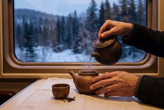 Hands of a man pouring hot water into a clay teapot to brew tea by a train window with a snowy forest outside