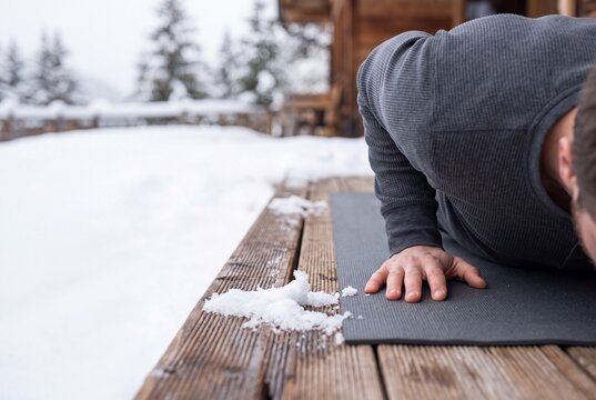 Fit man doing push ups on a yoga mat outdoors on a snowy wooden terrace during winter workout in the mountains