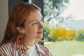 A young red-haired woman gazes thoughtfully out a window, smiling softly as natural sunlight highlights her face. Casual home setting with a blurred garden backdrop evokes calm, hope and quiet © Daniel