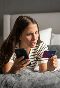 Frustrated woman lying on a bed, holding a smartphone and a credit card, looking stressed while encountering an issue with her online purchase or transaction
