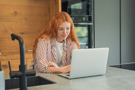 Young redhead woman focusing on her laptop while telecommuting from a modern home kitchen, embodying concepts of remote work, productivity, and flexible lifestyle