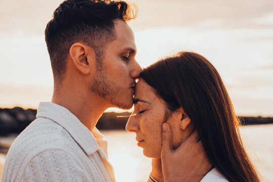 Young man tenderly kissing woman's forehead during golden hour, expressing deep connection, love, and romance while enjoying a serene moment together outdoors