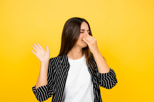 Young woman pinching her nose with a hand while waving another hand, expressing disgust and aversion to an unpleasant bad smell or stinky odor on a yellow background